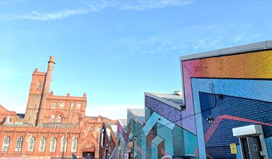 Cains Brewery Village From Outside. On the right is a jagged shaped building covered in colourful artwork. The red brick brewery building can be seen