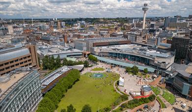 A drone shot of Chavasse Park in Liverpool ONE. A large green space amongst the built up shopping centre. The shot looks further towards the city and