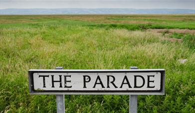 Road sign that says 'The Parade' next to some green Marshes with hills in the distance