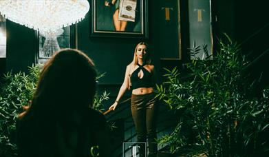 A girl wearing green trousers and a black cropped top is walking down the stairs in an ornate bar surrounded by plants