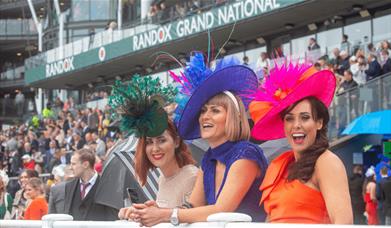 3 ladies dressed in large fascinator style hats in the stands at Aintree Racecourse. The signage in the stands behind reads 'Randox Grand National'