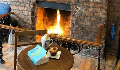 A roaring fire sits in an authentic, exposed-brick fire pit alcove. There are safety railings around the fire. In front of the fire sits a round table