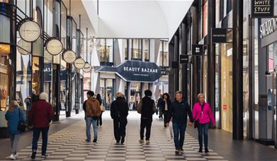 People walking through a selection of stores with large windows and black and white tiled floor. At the end of the corridor of stores you can see a la