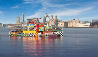 The Mersey Ferries brightly coloured Dazzle Ferry sailing on the Mersey River on a blue sky day. The iconic Liverpool skyline is behind the ferry.