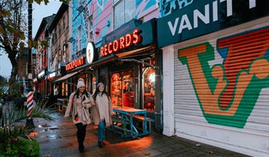 Two friends walk arm in arm along Victoria Road in New Brighton. They're next to a large, light up sign that reads 'Rockpoint Records' a bar and resta