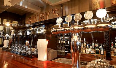 A traditional bar setup with real ale pumps and silver pumps. Behind the bar is wood panelled with 'RIGBY'S' in brass lettering.