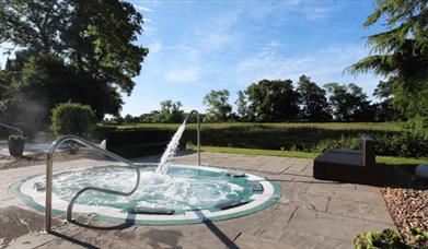 An outdoor sunken hot tub style pool with a large tap spraying water into the pool. Surrounding the pool is the countryside and lush grass.