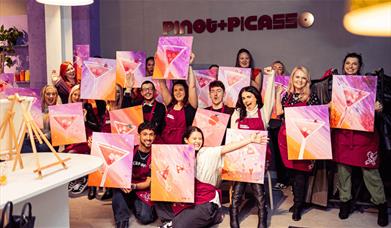 A group of people holding one canvas each, with a Martini glass painted on each. The canvas has pink, orange, white and red colours and the people hol